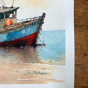 May include: A watercolor painting of a blue and red fishing boat resting on a sandy shore near calm water. The vessel features a cabin and visible rigging. The artist's signature, Joe Schaub, is written in dark ink on the lower sandy area.