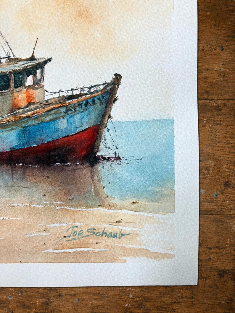 May include: A watercolor painting of a blue and red fishing boat resting on a sandy shore near calm water. The vessel features a cabin and visible rigging. The artist's signature, Joe Schaub, is written in dark ink on the lower sandy area.