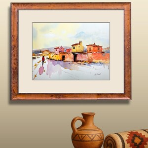 May include: Framed watercolour painting of a village scene. The artwork shows colourful buildings and a person walking on a path. The painting uses a warm colour palette with shades of orange, yellow, and red.