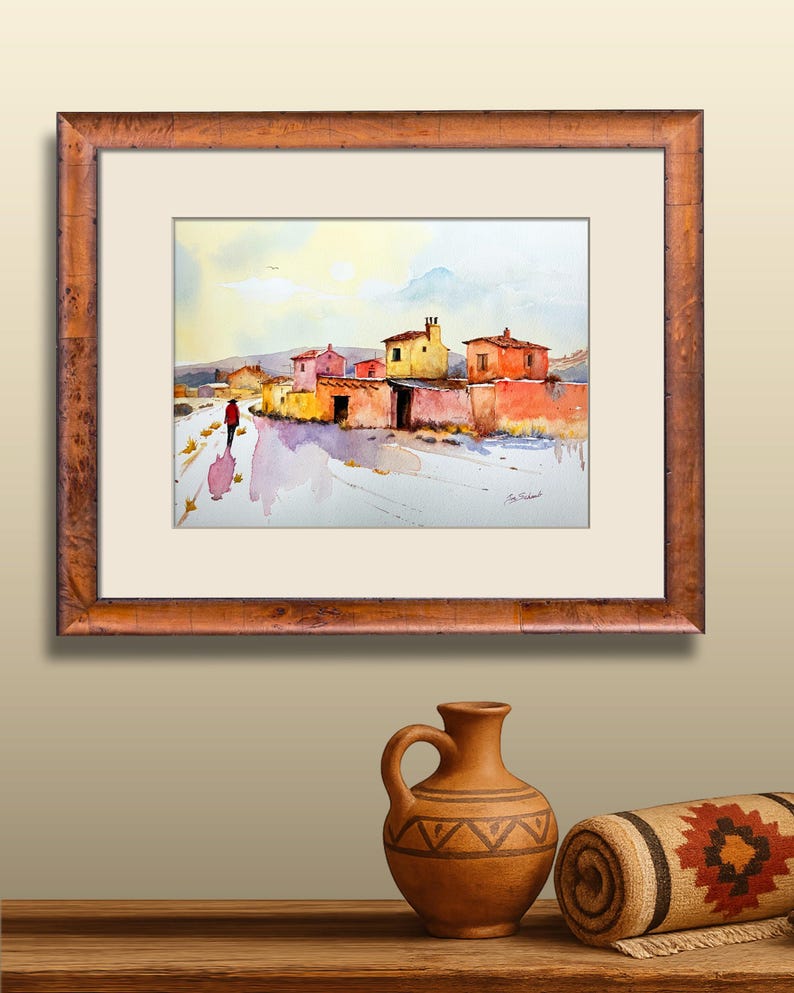 May include: Framed watercolour painting of a village scene. The artwork shows colourful buildings and a person walking on a path. The painting uses a warm colour palette with shades of orange, yellow, and red.