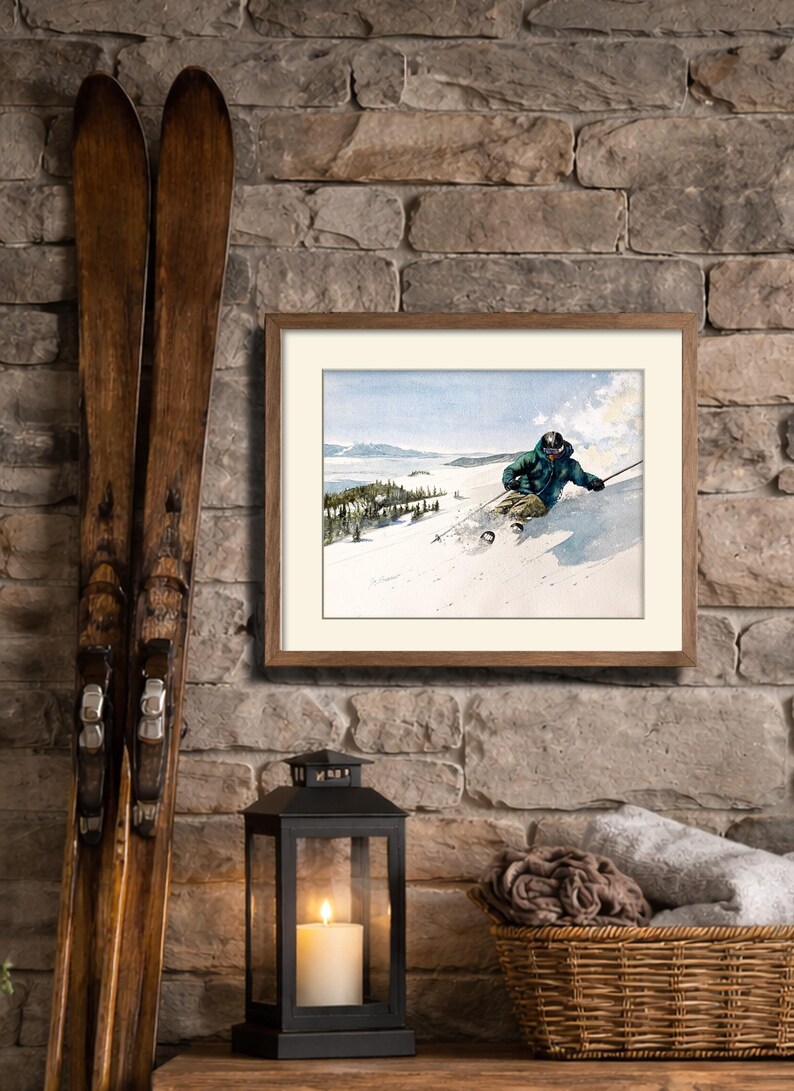 May include: A framed vintage watercolor print of a skier on a snowy slope, with a wooden frame. Two antique skis lean against a stone wall. A black lantern with a lit candle and a wicker basket with rolled blankets complete the scene.