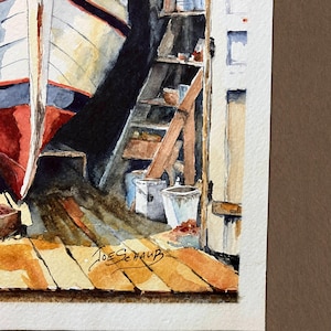 May include: A watercolor painting depicting a boat hull in a workshop with wooden floorboards, shelves holding various containers, and a white door frame. The artist's signature, Joe Schaub, is visible in black ink on the lower wooden floor area.