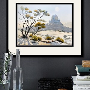 May include: Watercolor landscape art print featuring a desert scene with a prominent mesa, trees, and shrubs. The artwork is framed in black and matted with a white border. The print is displayed on a white shelf with decorative items.