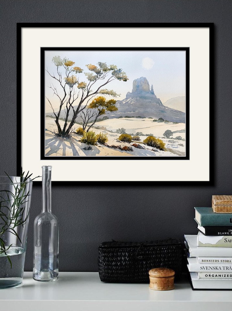 May include: Watercolor landscape art print featuring a desert scene with a prominent mesa, trees, and shrubs. The artwork is framed in black and matted with a white border. The print is displayed on a white shelf with decorative items.
