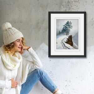 K&ouml;nnte beinhalten: Ein gerahmtes Aquarellgem&auml;lde zeigt eine schneebedeckte Klippe mit immergr&uuml;nen B&auml;umen. Das Kunstwerk befindet sich in einem schwarzen Rahmen mit wei&szlig;em Passepartout. Das Gem&auml;lde wird an einer grauen Wand ausgestellt. Eine Person tr&auml;gt eine wei&szlig;e Strickm&uuml;tze, einen Schal und einen Pullover.
