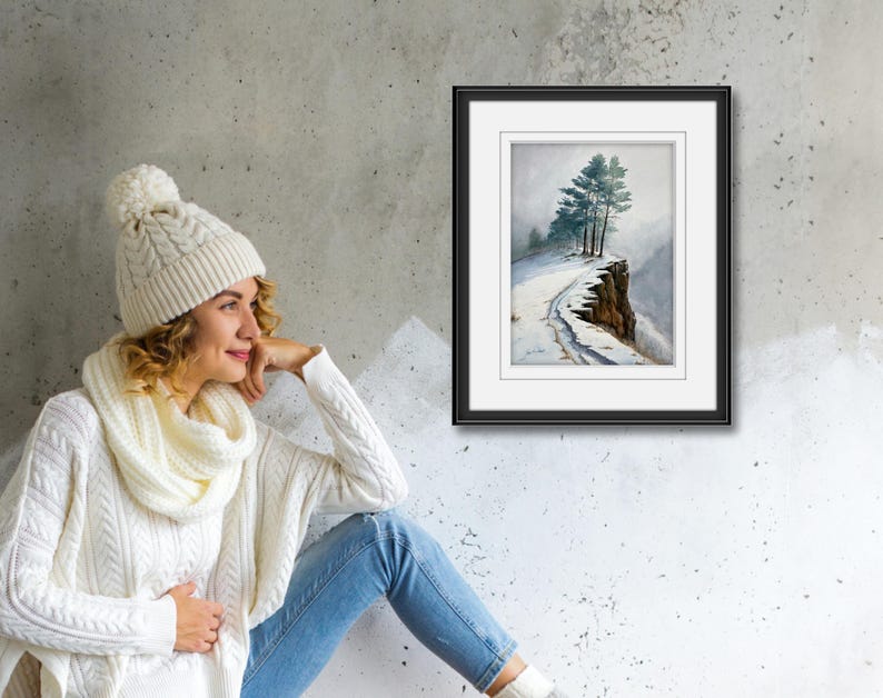 K&ouml;nnte beinhalten: Ein gerahmtes Aquarellgem&auml;lde zeigt eine schneebedeckte Klippe mit immergr&uuml;nen B&auml;umen. Das Kunstwerk befindet sich in einem schwarzen Rahmen mit wei&szlig;em Passepartout. Das Gem&auml;lde wird an einer grauen Wand ausgestellt. Eine Person tr&auml;gt eine wei&szlig;e Strickm&uuml;tze, einen Schal und einen Pullover.