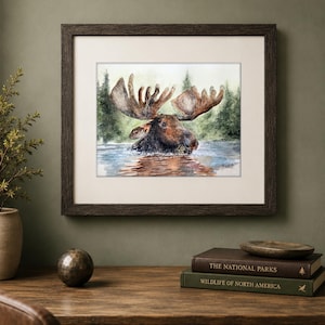 May include: A framed watercolor painting depicting a brown moose wading in water with a forest background. The artwork features a signature in the bottom right corner and is enclosed in a dark brown wooden frame with a white matting border.
