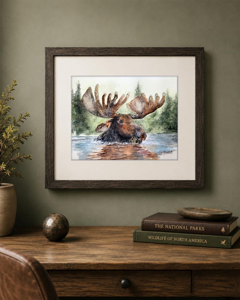 May include: A framed watercolor painting depicting a brown moose wading in water with a forest background. The artwork features a signature in the bottom right corner and is enclosed in a dark brown wooden frame with a white matting border.