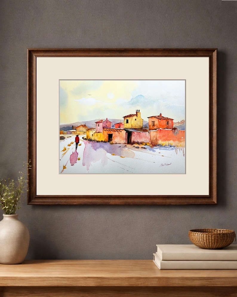 May include: Watercolour painting depicting a village scene with vibrant buildings and a person in a red coat walking along a path. The artwork is framed in a dark brown wooden frame with a white mount.