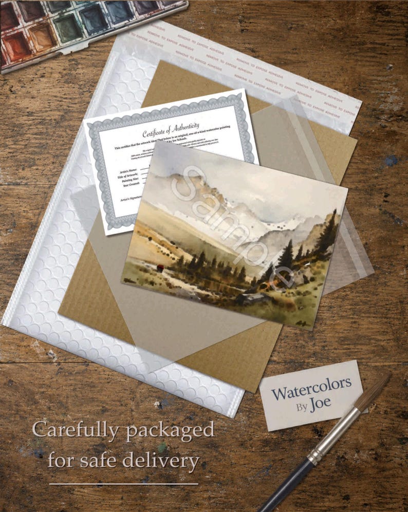 May include: A watercolor landscape painting of mountains and pine trees, accompanied by a Certificate of Authenticity, a Watercolors by Joe business card, and a paintbrush, all arranged on a wooden surface with packaging materials.