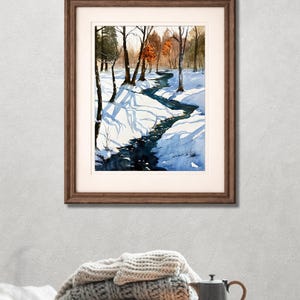 May include: Watercolor painting of a winter landscape with a winding stream. The artwork, framed in a brown wooden frame, depicts snow-covered ground, bare trees, and a dark blue stream. The painting is displayed on a wall above a stack of blankets and a coffee table.