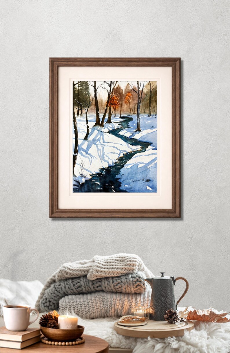 May include: Watercolor painting of a winter landscape with a winding stream. The artwork, framed in a brown wooden frame, depicts snow-covered ground, bare trees, and a dark blue stream. The painting is displayed on a wall above a stack of blankets and a coffee table.