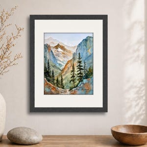 May include: Watercolor painting of a mountain landscape in a black frame. The artwork depicts a valley with tall pine trees, rocky cliffs, and snow-capped mountains in the distance. The color palette includes blues, greens, and browns.