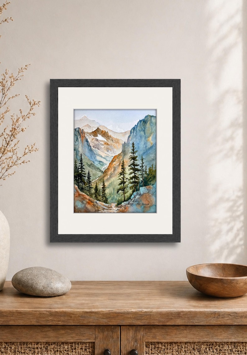 May include: Watercolor painting of a mountain landscape in a black frame. The artwork depicts a valley with tall pine trees, rocky cliffs, and snow-capped mountains in the distance. The color palette includes blues, greens, and browns.