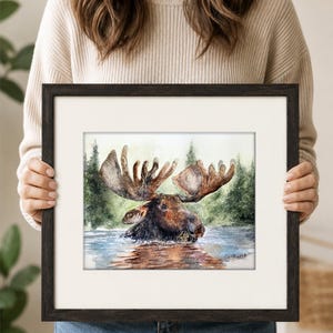 May include: A framed watercolor painting of a brown moose with large antlers wading in water, set against a backdrop of green pine trees. The artwork features a white mat and a dark brown wooden frame, with a signature visible in the bottom right.