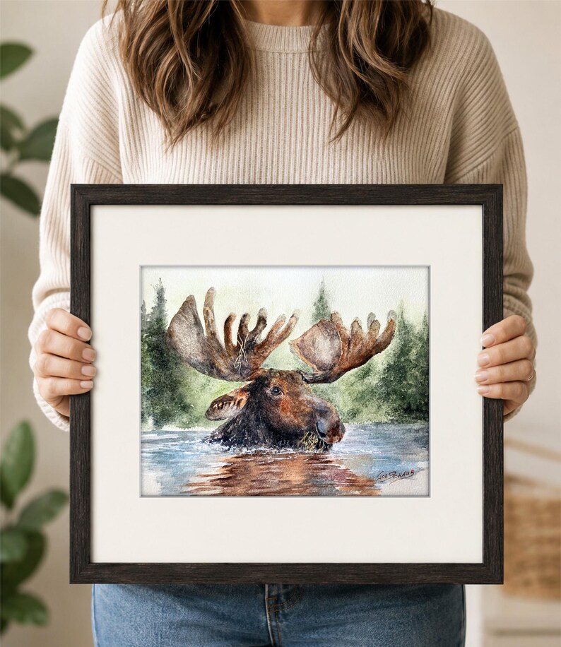 May include: A framed watercolor painting of a brown moose with large antlers wading in water, set against a backdrop of green pine trees. The artwork features a white mat and a dark brown wooden frame, with a signature visible in the bottom right.