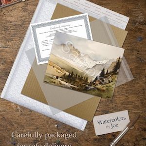 May include: A watercolor painting of a mountain landscape with pine trees and a stream, accompanied by a Certificate of Authenticity, a business card reading Watercolors By Joe, and a paintbrush, all arranged on a wooden surface.