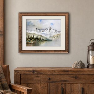May include: A framed watercolor painting depicting a snow-capped mountain range rising above a calm lake. The landscape features dark green pine trees along the shoreline, soft blue and grey sky tones, and a textured brown wooden frame with a white mat.