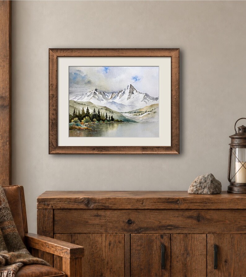 May include: A framed watercolor painting depicting a snow-capped mountain range rising above a calm lake. The landscape features dark green pine trees along the shoreline, soft blue and grey sky tones, and a textured brown wooden frame with a white mat.