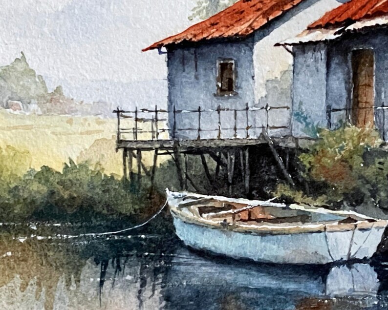 May include: Watercolor painting of a coastal scene featuring two weathered, light blue buildings with red roofs. A small white boat floats in the water, tied to the shore. The artwork depicts a serene landscape with muted colors and a sense of tranquility.
