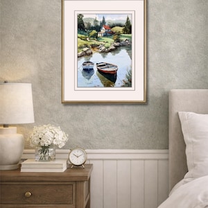 May include: Framed watercolor painting of a serene lakeside scene. Two small boats float on the water, reflecting the sky and surrounding trees. A house with a red roof sits on the shore, framed by a gold-colored border.
