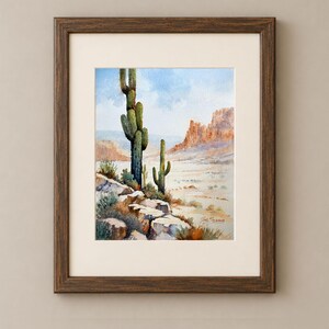 May include: A framed watercolor painting depicting a desert landscape with two tall saguaro cacti in the foreground, rocky terrain, and distant mesas under a light blue sky. The artwork is enclosed in a dark brown wooden frame with a cream mat.