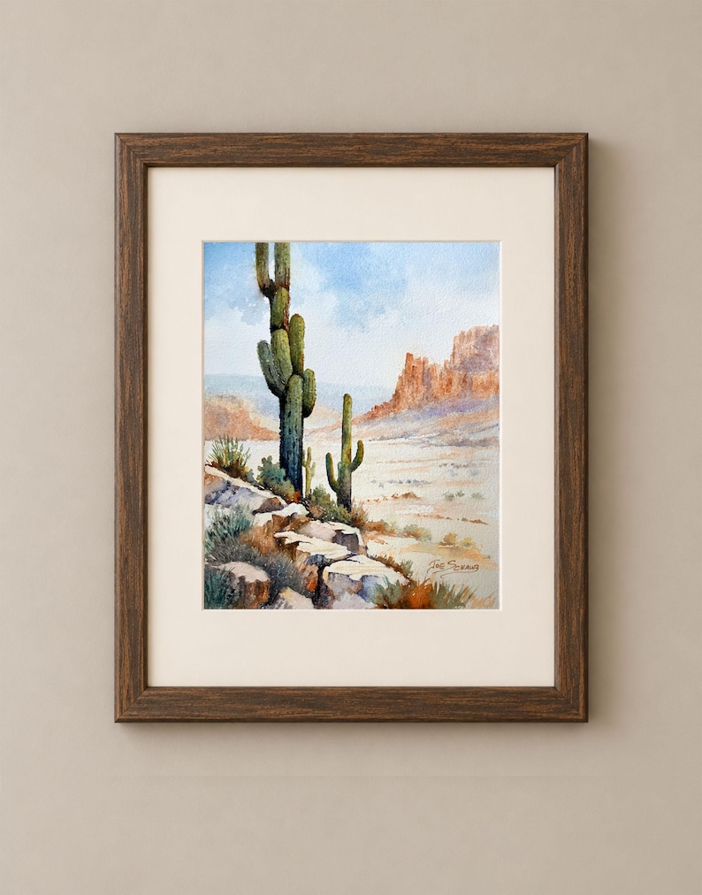 May include: A framed watercolor painting depicting a desert landscape with two tall saguaro cacti in the foreground, rocky terrain, and distant mesas under a light blue sky. The artwork is enclosed in a dark brown wooden frame with a cream mat.