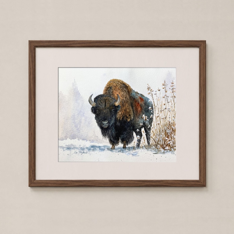 May include: A framed watercolor painting of a bison standing in a snowy landscape with dry grasses. The bison has dark brown and black fur with a thick mane. The artwork is enclosed in a dark brown wooden frame with a light-colored mat border.