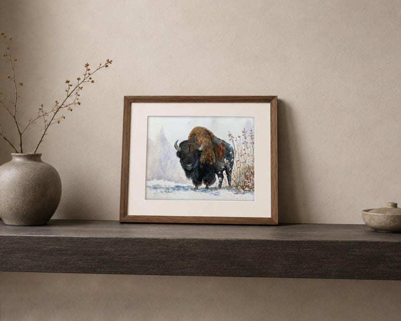 May include: A framed art print featuring a watercolor illustration of a bison standing in a snowy landscape with dry grasses. The artwork is enclosed in a rectangular wooden frame with a white mat border, showcasing the animal's dark fur and texture.