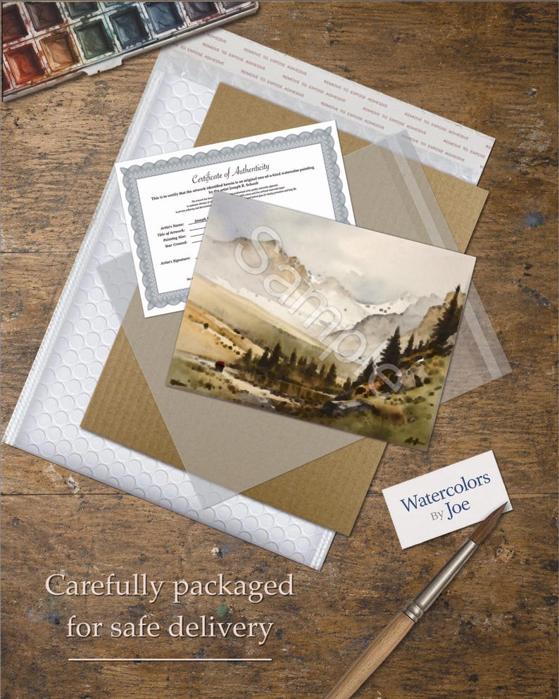 May include: A watercolor landscape painting of mountains and pine trees, a Certificate of Authenticity, a white bubble mailer, a brown cardboard insert, a business card reading Watercolors By Joe, and a wooden paintbrush on a wooden surface.