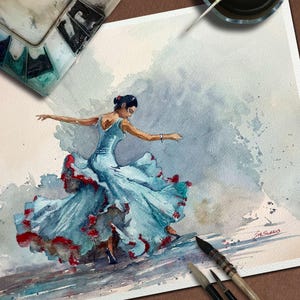 May include: Watercolor painting of a flamenco dancer in a flowing blue dress with red accents. The artwork captures the dancer mid-motion, with arms outstretched. Paintbrushes, a palette, and ink are visible, suggesting an artist's workspace.