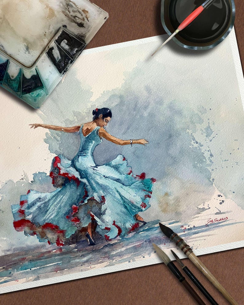 May include: Watercolor painting of a flamenco dancer in a flowing blue dress with red accents. The artwork captures the dancer mid-motion, with arms outstretched. Paintbrushes, a palette, and ink are visible, suggesting an artist's workspace.