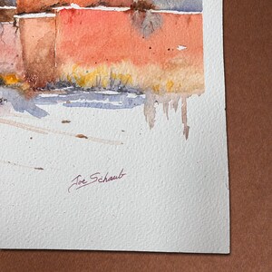 May include: Watercolour painting depicting a building with red-orange walls and small windows. The artwork uses a muted colour palette with shades of red, brown, and blue. The artist's signature, "Joe Schaub," is visible in the lower left corner.