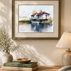 May include: Watercolor painting of two small white houses with red roofs on stilts over water. A small white boat is docked nearby. The artwork is framed in a wooden frame and displayed on a wooden table with books and a lamp.