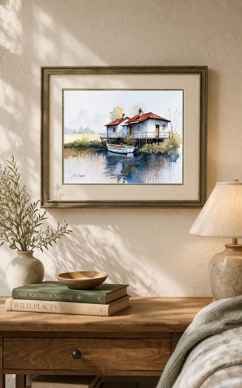 May include: Watercolor painting of two small white houses with red roofs on stilts over water. A small white boat is docked nearby. The artwork is framed in a wooden frame and displayed on a wooden table with books and a lamp.
