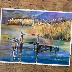 Puede incluir: Una pintura de acuarela sobre papel texturizado que muestra un muelle de madera extendi&eacute;ndose en aguas tranquilas con tres aves posadas en los postes. El fondo presenta juncos dorados altos bajo un cielo azul y violeta. Firma Joe Schaub visible.
