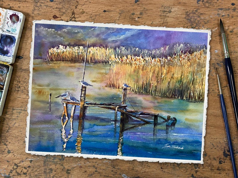 Puede incluir: Una pintura de acuarela sobre papel texturizado que muestra un muelle de madera extendi&eacute;ndose en aguas tranquilas con tres aves posadas en los postes. El fondo presenta juncos dorados altos bajo un cielo azul y violeta. Firma Joe Schaub visible.