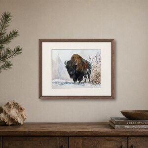 May include: A framed horizontal art print featuring a watercolor painting of a dark brown bison standing in a snowy landscape with sparse vegetation. The artwork is enclosed in a dark wood frame with a wide off-white mat border.
