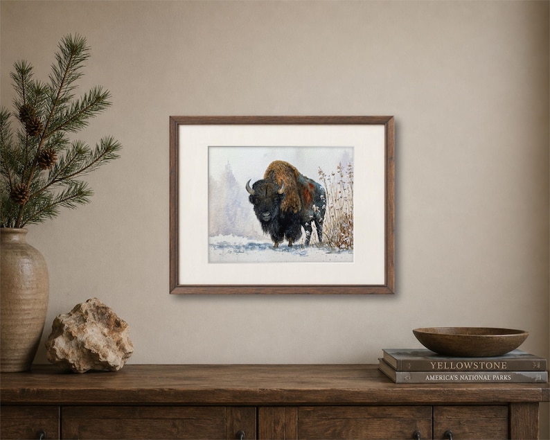 May include: A framed horizontal art print featuring a watercolor painting of a dark brown bison standing in a snowy landscape with sparse vegetation. The artwork is enclosed in a dark wood frame with a wide off-white mat border.