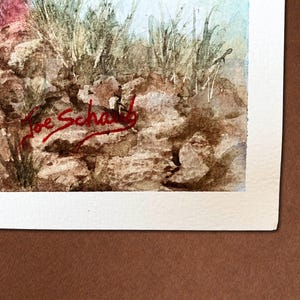 Op de afbeelding: Aquarel schilderij van een landschap met een rotsachtige voorgrond, hoog groen gras en een lichtblauwe lucht. De handtekening van de kunstenaar, "Joe Schwab", staat met rode inkt in de linker benedenhoek. Het kunstwerk staat op wit papier.