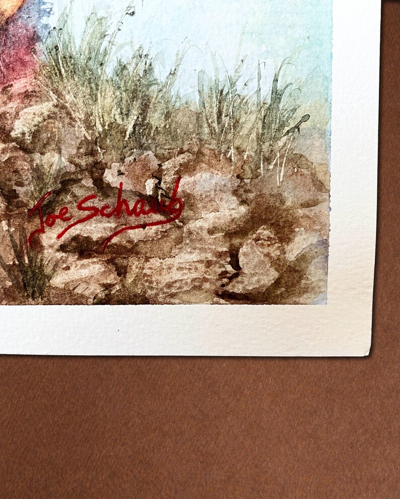 Op de afbeelding: Aquarel schilderij van een landschap met een rotsachtige voorgrond, hoog groen gras en een lichtblauwe lucht. De handtekening van de kunstenaar, "Joe Schwab", staat met rode inkt in de linker benedenhoek. Het kunstwerk staat op wit papier.