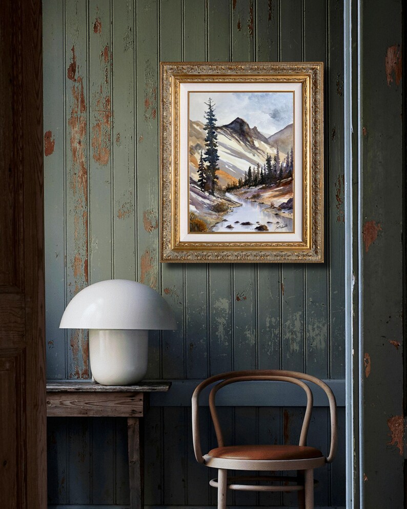 May include: A framed landscape painting with a gold ornate frame hangs on a distressed green wall. The painting depicts a mountain scene with a river and trees. A white mushroom-shaped lamp sits on a wooden table below the painting.