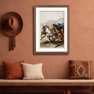 May include: A framed watercolor painting depicts cowboys on horseback roping cattle. The artwork is in a wooden frame and hangs on a terracotta-colored wall. A brown hat and pillows are also visible.