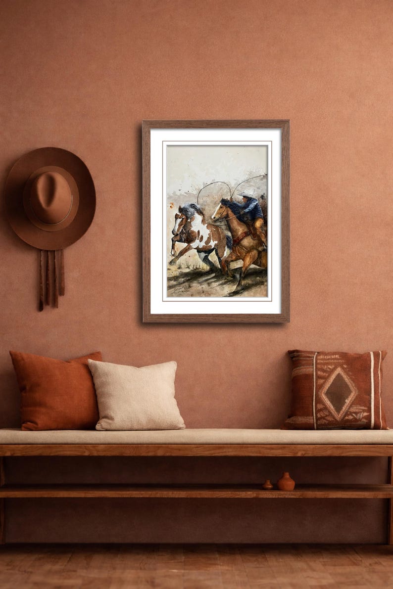 May include: A framed watercolor painting depicts cowboys on horseback roping cattle. The artwork is in a wooden frame and hangs on a terracotta-colored wall. A brown hat and pillows are also visible.