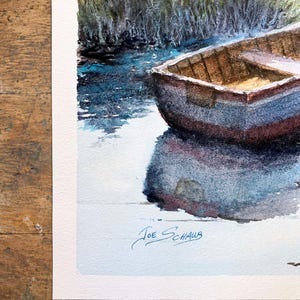 Pu&ograve; includere: Un dipinto ad acquerello che raffigura una piccola barca di legno che galleggia su un'acqua blu scuro vicino a una riva erbosa. La barca proietta un profondo riflesso sulla superficie dell'acqua. La firma Joe Schaub &egrave; scritta in basso a sinistra.