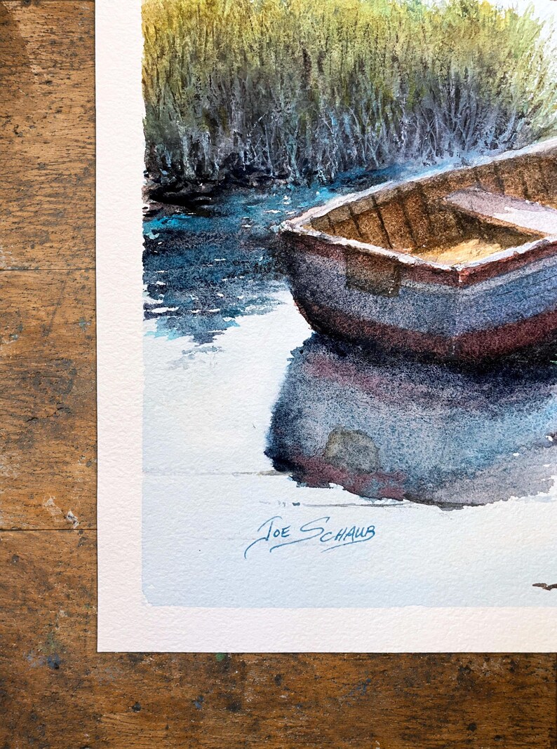 Pu&ograve; includere: Un dipinto ad acquerello che raffigura una piccola barca di legno che galleggia su un'acqua blu scuro vicino a una riva erbosa. La barca proietta un profondo riflesso sulla superficie dell'acqua. La firma Joe Schaub &egrave; scritta in basso a sinistra.
