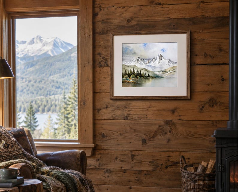 May include: A framed watercolor painting depicting a snow-capped mountain range above a calm lake and a line of dark green pine trees. The artwork is displayed in a light-colored mat and a brown wooden frame, hanging on a rustic wood-paneled wall.