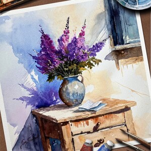 May include: Watercolor painting of a still life featuring a bouquet of purple flowers in a blue vase on a wooden table. A small book rests on the table, with paint tubes and brushes nearby. The artwork has a soft, natural light.