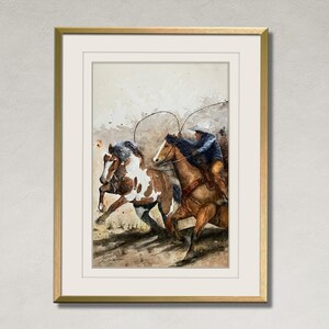 May include: Watercolor painting of a cowboy on horseback roping a horse. The painting features a brown and white horse and a brown horse with a cowboy wearing a blue jacket and a white hat. The artwork is framed in a gold frame.