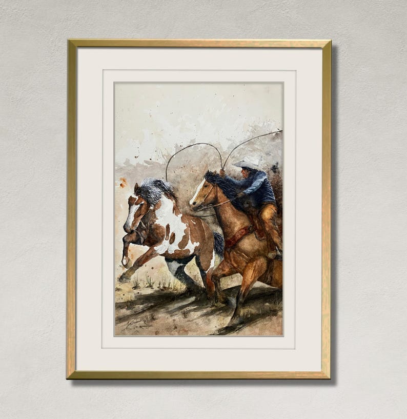 May include: Watercolor painting of a cowboy on horseback roping a horse. The painting features a brown and white horse and a brown horse with a cowboy wearing a blue jacket and a white hat. The artwork is framed in a gold frame.